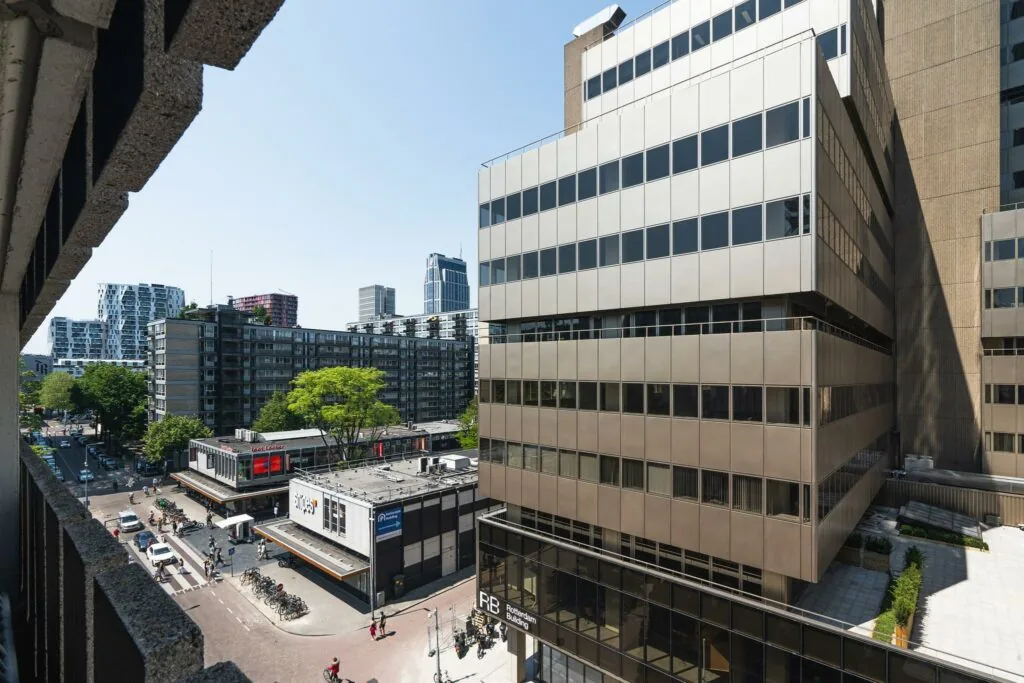 Uitzicht op de Aert van Nesstraat in Rotterdam met moderne kantoorgebouwen, winkels en fietsende voorbijgangers op een zonnige dag.